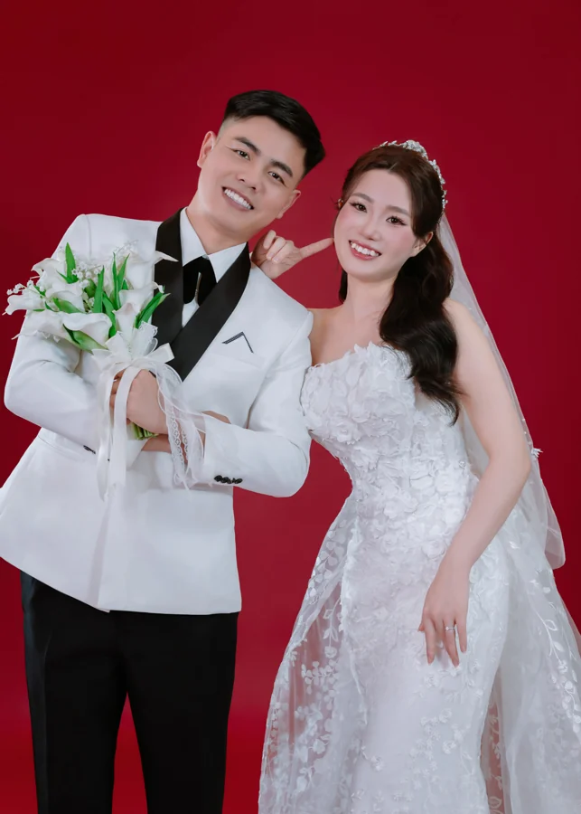 Cặp đôi cười tươi trong trang phục cưới trên nền studio đỏ